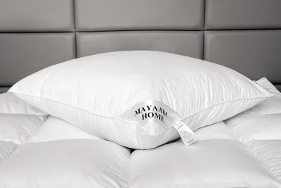 A white pillow with a brand tag is placed on top of other white bedding, with a grey upholstered headboard in the background.