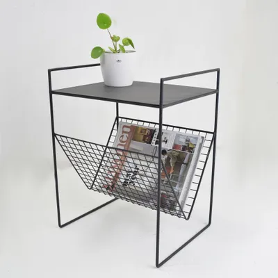 A black metal side table with a magazine rack and a potted plant on top, set against a white background.
