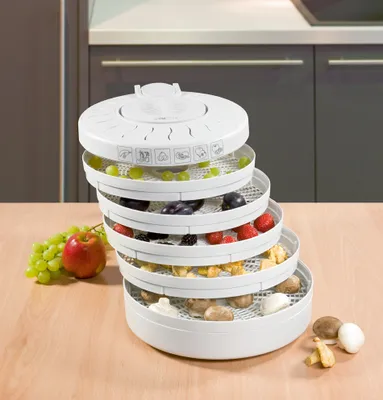 A white food dehydrator with multiple stacked trays filled with various fruits and vegetables, placed on a wooden table. A bunch of green grapes and a red apple are on the left side of the dehydrator.