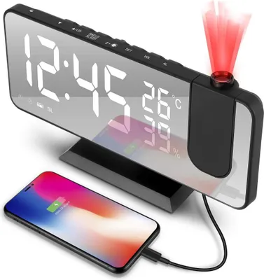 A digital alarm clock with a projected display and a smartphone charging on its base.