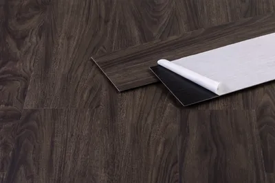 A close-up shot of two vinyl flooring planks on a dark wood-like surface. One plank is fully visible, while the other is partially peeled back, revealing its adhesive backing.