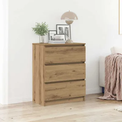 A wooden dresser with three drawers is placed against a white wall in a room. There are decorative items on top of the dresser, and a pink blanket is draped over a sofa in the background.