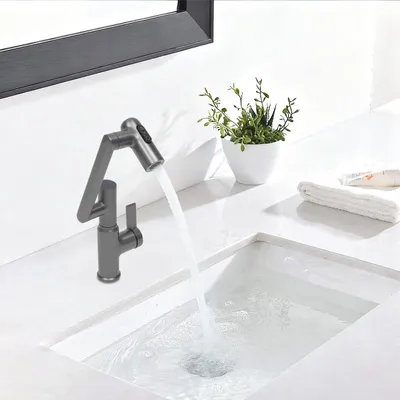 A modern bathroom or kitchen sink with a gray faucet running water into the sink. A small potted plant and a folded towel are on the counter.