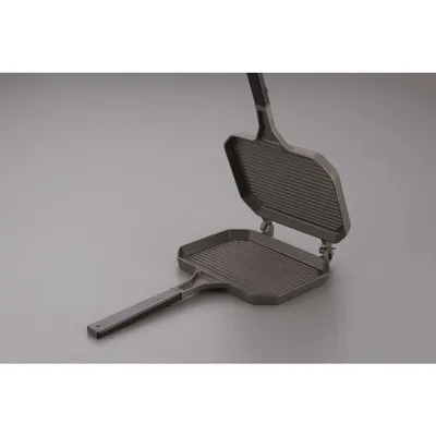 A double-sided sandwich toaster with a ribbed surface is shown open against a plain gray background.