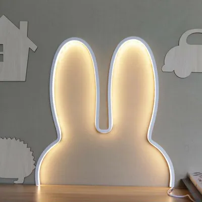 A bunny ear-shaped night light is mounted on a gray wall above a wooden surface. Other wooden cutouts of a house, a car, and a hedgehog are also visible on the wall.