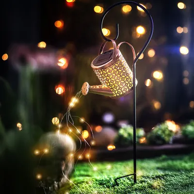 A decorative watering can with lights is displayed in a garden at night.