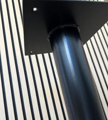A close-up of a black metal table base with a square mounting plate, set against a background of light-colored wooden slats with dark vertical stripes.