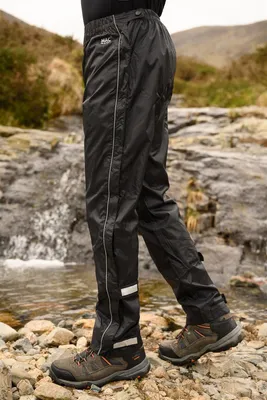 A person wearing black rain pants and hiking boots stands on rocks near a waterfall.