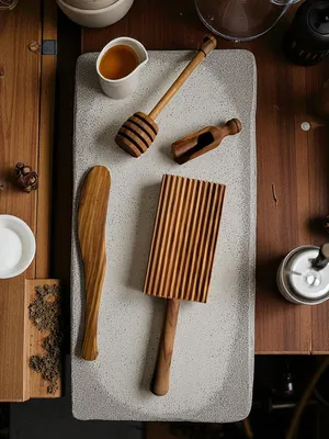 A collection of wooden kitchen utensils, including a gnocchi board, honey dipper, and scoop, arranged on a light gray textured tray.