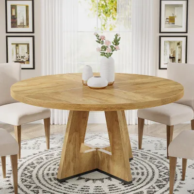 A round wooden dining table with chairs around it, set in a well-lit room with artwork on the walls and a decorative rug on the floor.