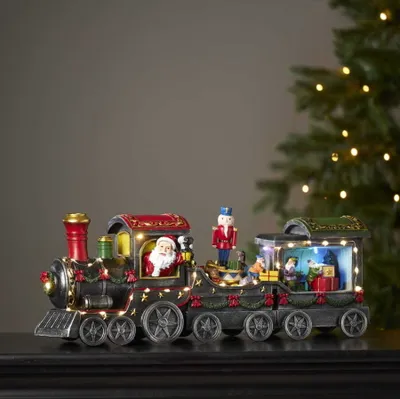 A festive Christmas train decoration with Santa Claus and nutcracker figures, illuminated by small LED lights, placed on a dark surface with a blurred Christmas tree in the background.