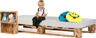 A young child sits on a bed frame made of wooden pallets, with decorative pillows and wooden crates nearby.