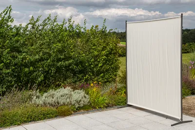 A beige textile privacy screen stands on a paved patio next to a garden with lush green bushes and plants.