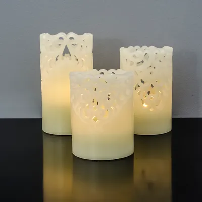 Three LED candles with intricate cut-out patterns are displayed on a reflective black surface against a gray wall.