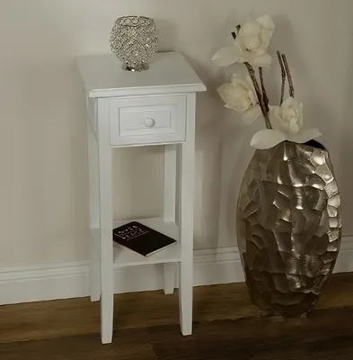 A white telephone table with a drawer and a shelf, placed against a wall with a large decorative vase and flowers next to it.