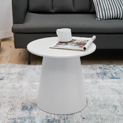 A white round side table with a magazine and a mug on it, placed on a rug in front of a sofa.