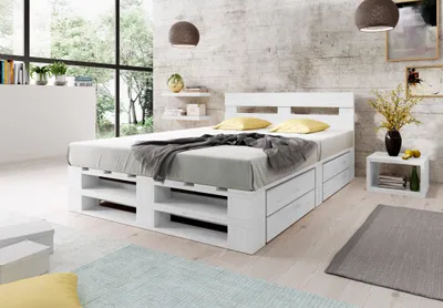 A bedroom scene featuring a white pallet bed with storage drawers, set against a textured wall and large windows.