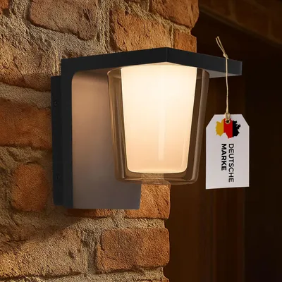 A modern LED outdoor wall light mounted on a brick wall, with a tag indicating it is a German brand.