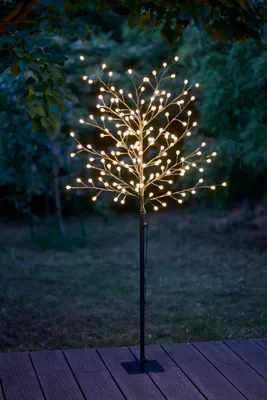 An LED tree light with warm white LEDs is displayed outdoors at dusk, standing on a wooden deck with a garden in the background.