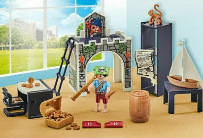 A Playmobil pirate-themed playset is arranged on a sandy surface, with a window and outdoor scene in the background.