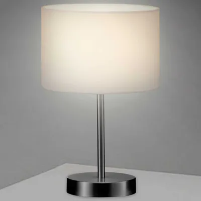 A table lamp with a white cylindrical shade and a silver metallic base and stem, placed on a white surface against a light gray background.