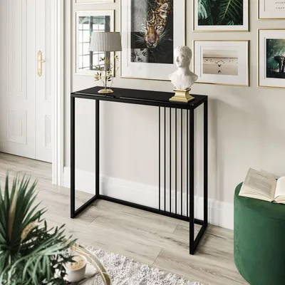 A modern console table with a black glass top and a black metal frame, decorated with vertical metal bars on one side. It is placed in a well-lit room with framed artwork on the wall and a green ottoman nearby.