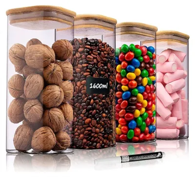 Vier klare Glasvorratsbehälter mit quadratischen Bambusdeckeln, gefüllt mit Walnüssen, Kaffeebohnen, bunten Bonbons und rosa Marshmallows, mit einem weißen Kreidestift davor