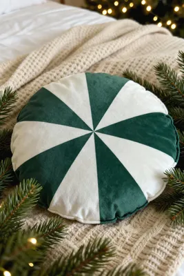 A round pillow with green and white stripes is placed on a cream-colored knitted blanket, surrounded by pine branches and fairy lights.