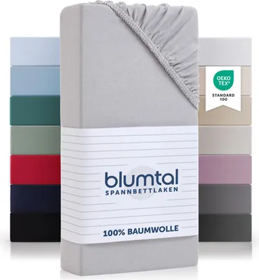 A stack of folded fitted sheets in various colors, with a close-up of a grey fitted sheet in the foreground. The grey sheet has a label with brand and product information.