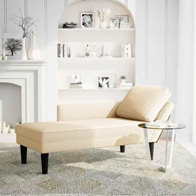 A cream-colored chaise lounge with a pillow sits next to a small glass-top side table in a well-lit room with shelves in the background.
