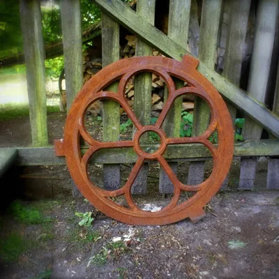A rusty metal decorative wheel is leaning against a wooden fence in an outdoor setting.