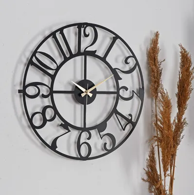 A black metal wall clock with Roman numerals is mounted on a light gray wall, with dried grass stalks to the right.
