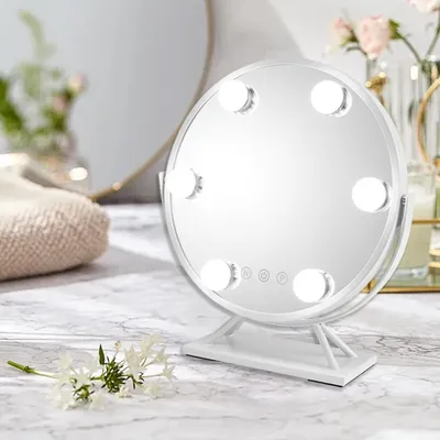 A white makeup mirror with LED lights sits on a marble countertop, with a blurred background of a larger mirror and flowers.