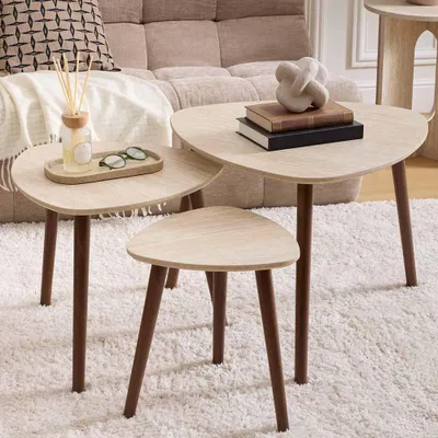 A set of three nesting tables with a light wood grain finish and dark brown legs, arranged on a fluffy white rug in a living room setting. Decorative items like books, glasses, and a diffuser are placed on the tables.