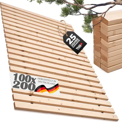 A wooden slatted base for a bed, viewed from an angle with a stack of similar bases in the background and a branch with green leaves overhead.