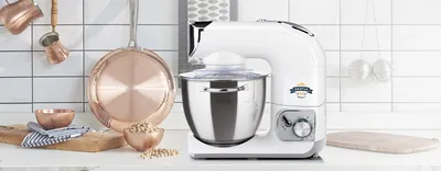 A white kitchen mixer is displayed on a countertop with copper kitchenware and ingredients in the background.