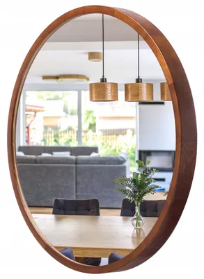 A round wooden mirror reflects a dining area with chairs and a table, and a living room with a sofa and fireplace.
