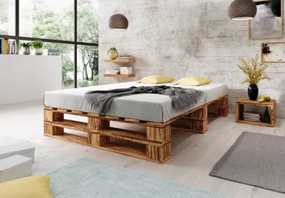 A bedroom with a pallet bed, pillows, and a blanket, set against a textured wall and large windows.