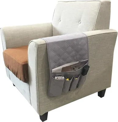 A close-up shot of a gray fabric organizer attached to the armrest of a beige armchair. The organizer has multiple pockets holding items like a remote control and magazines.