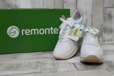 A pair of white Remonte sneakers are displayed next to their green packaging box on a wooden floor.