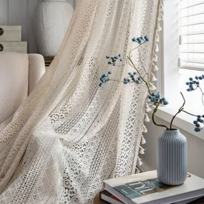 A close-up of a lace curtain with tassels, next to a vase with blue berries and a stack of books.