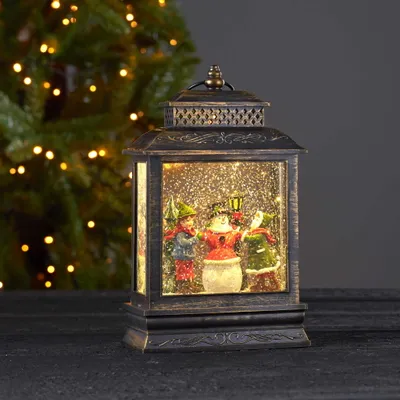 A decorative LED lantern with a Christmas scene inside, set against a blurred Christmas tree background.