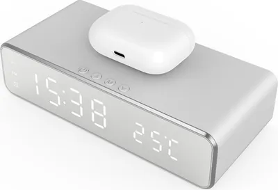 A silver digital alarm clock with a white wireless charging case on top, displaying the time and temperature.