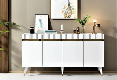 A white sideboard with gold accents is displayed in a room setting, decorated with artwork and a lamp.