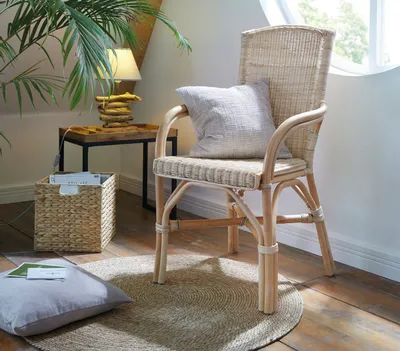 A rattan chair with a cushion is placed next to a side table with a lamp and a basket, in a room with natural light.