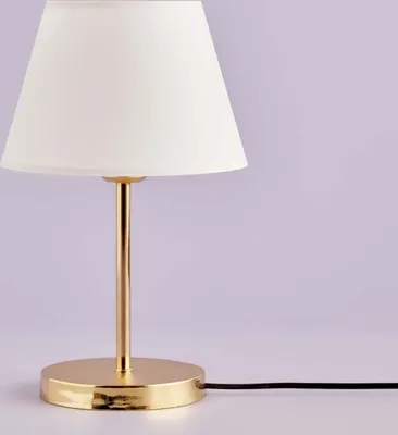 A close-up studio shot of a white table lamp with a gold base and stem, set against a light purple and white background.