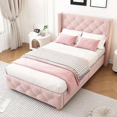 A pink upholstered bed with a tufted headboard and footboard, dressed with white and pink bedding and a gray and white throw blanket, is shown in a bedroom setting.