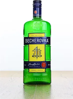 A green glass bottle of Becherovka herbal liqueur stands on a wooden surface against a white background.