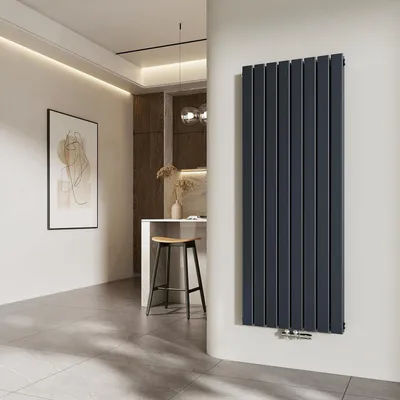A modern interior with a vertical radiator as the main focus, set against a white wall. The background shows a glimpse of a kitchen area with a stool and artwork on the wall.
