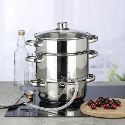 A stainless steel steamer with a glass lid and a transparent tube, placed on a black induction hob. Two glass bottles and a pile of cherries are visible in the background and foreground.